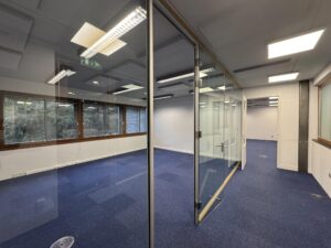 Modern empty office space on De Ruijterkade with blue carpet, glass partition walls, and large windows.